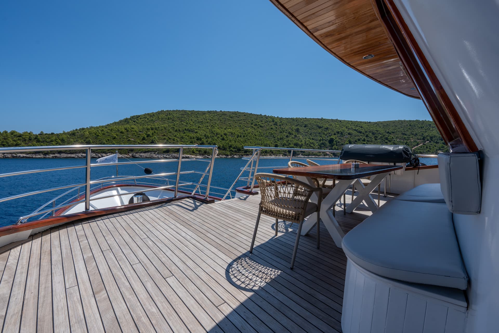 Yacht deck with wooden dining table and chairs overlooking calm blue coastal waters and green hills
