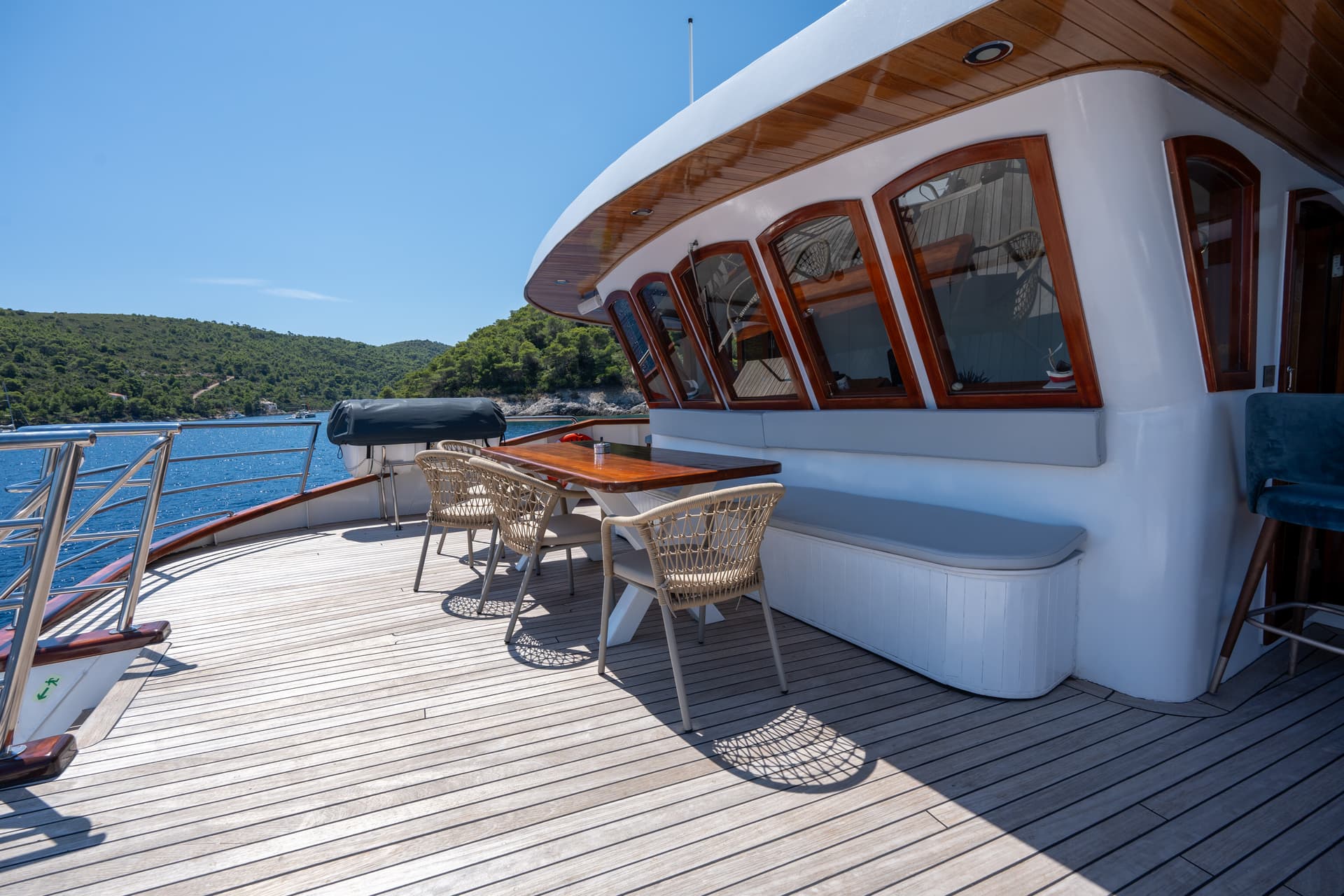 Wooden deck of yacht with table, chairs, and seating area overlooking blue coastal waters