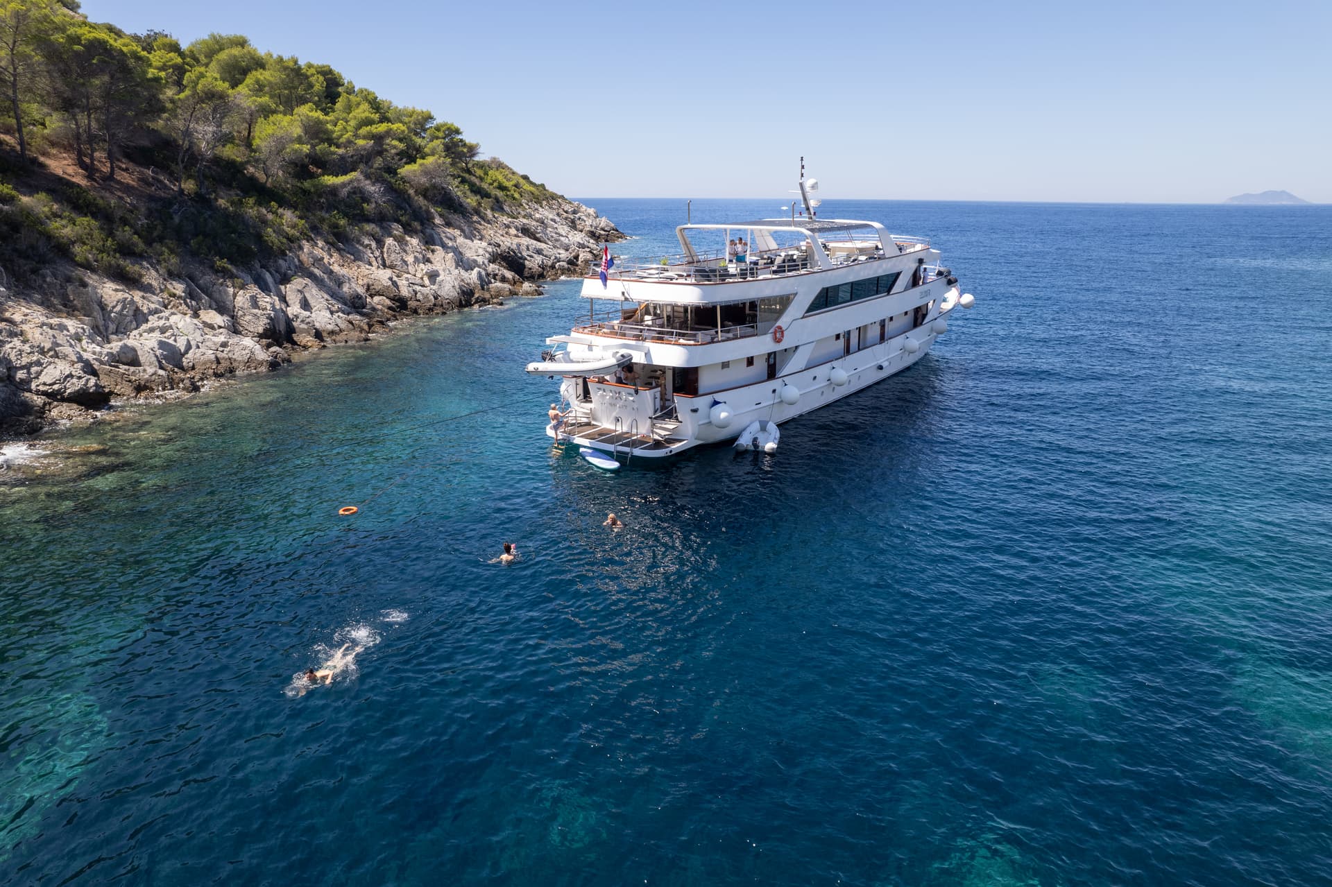 Large white yacht anchored in clear blue coastal waters with people swimming near a rocky shore