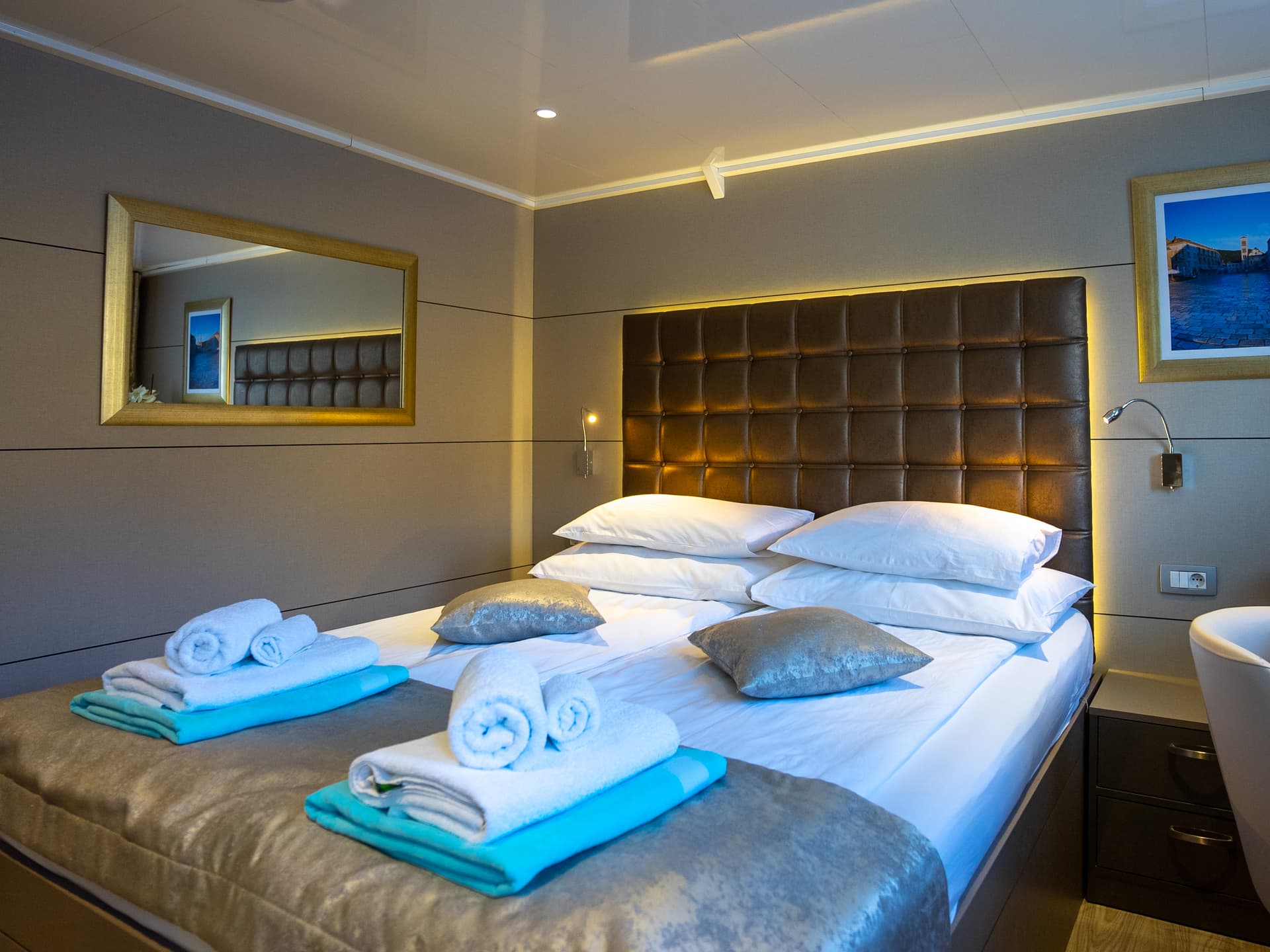 Cabin bedroom with a made bed, folded towels, and a brown tufted headboard on the main deck of a ship
