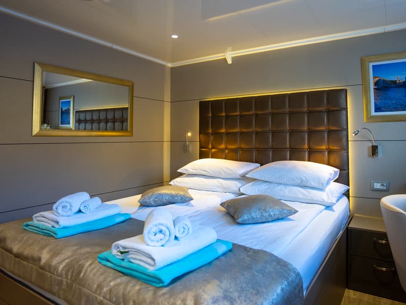 Cabin bedroom with a made bed, folded towels, and a brown tufted headboard on the main deck of a ship