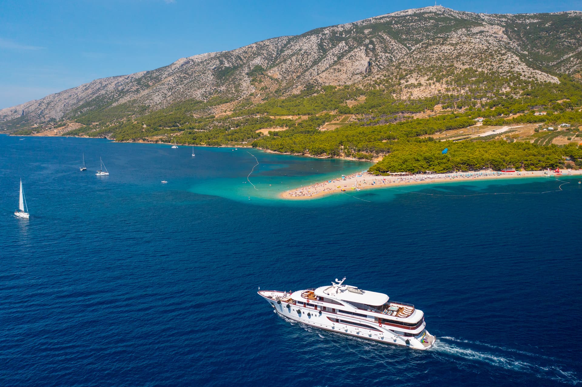 Large white yacht cruising on blue coastal waters with a beach and mountain backdrop