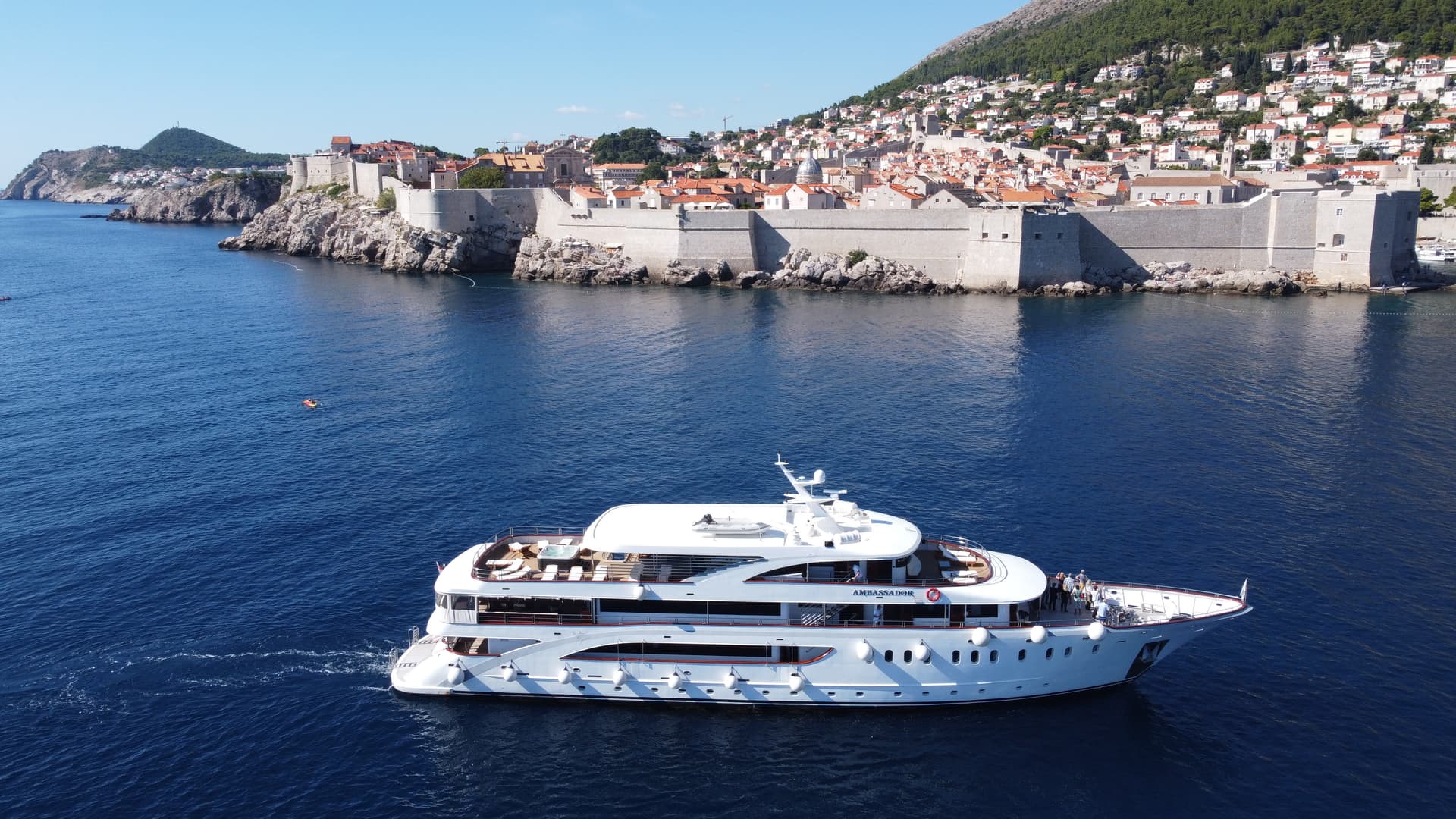 Large white yacht cruising on blue sea near historic walled coastal city with orange roofs
