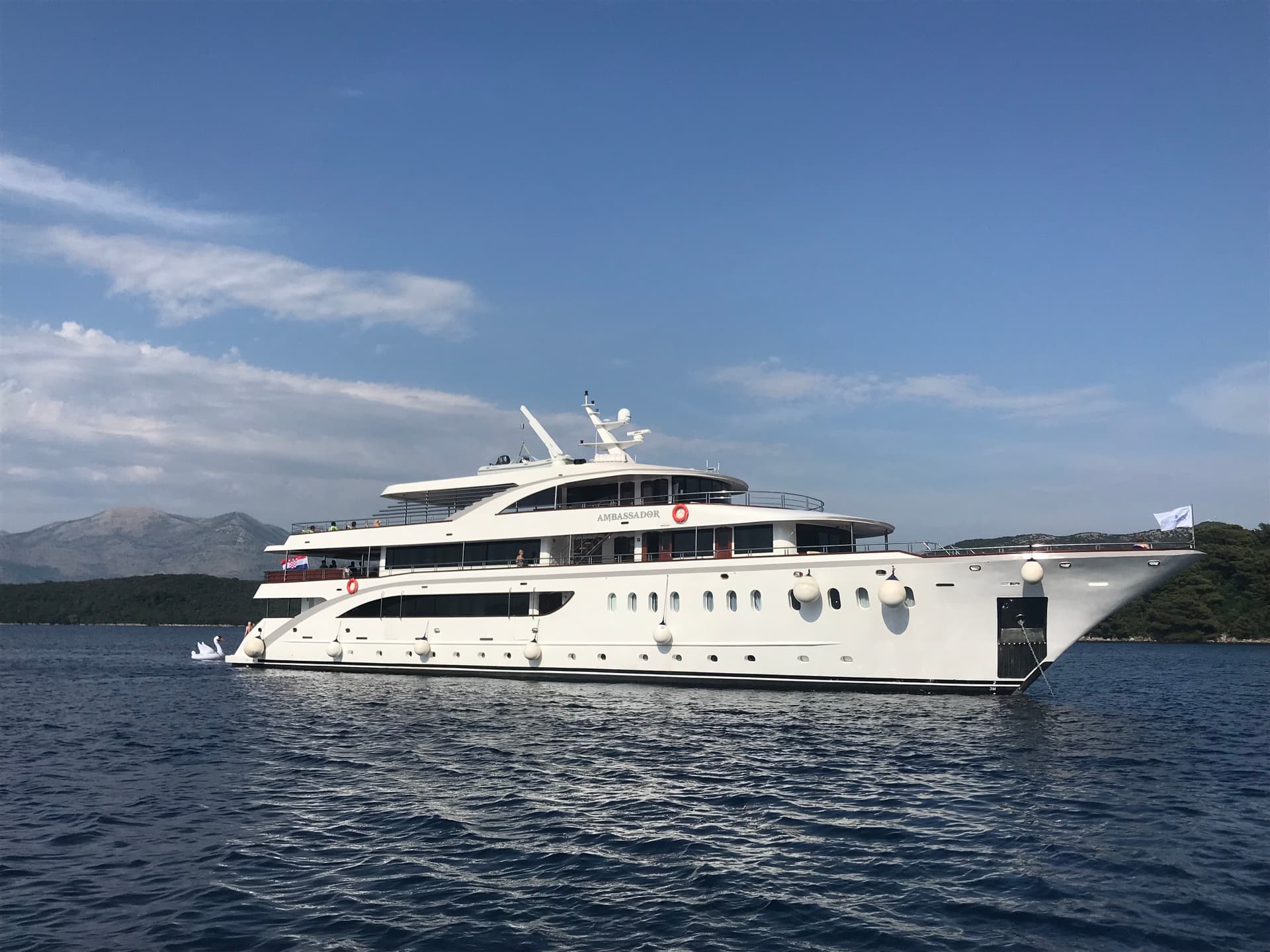 Large white yacht named Ambassador anchored on calm blue water with distant hills
