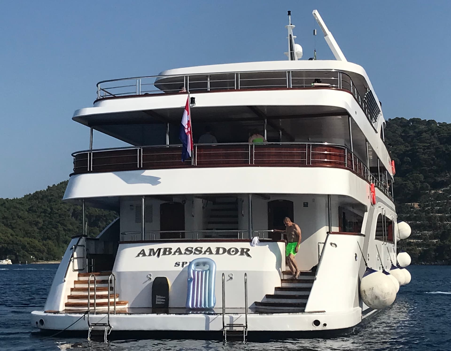 Large white yacht named Ambassador on calm blue coastal waters with green forested hills in the background