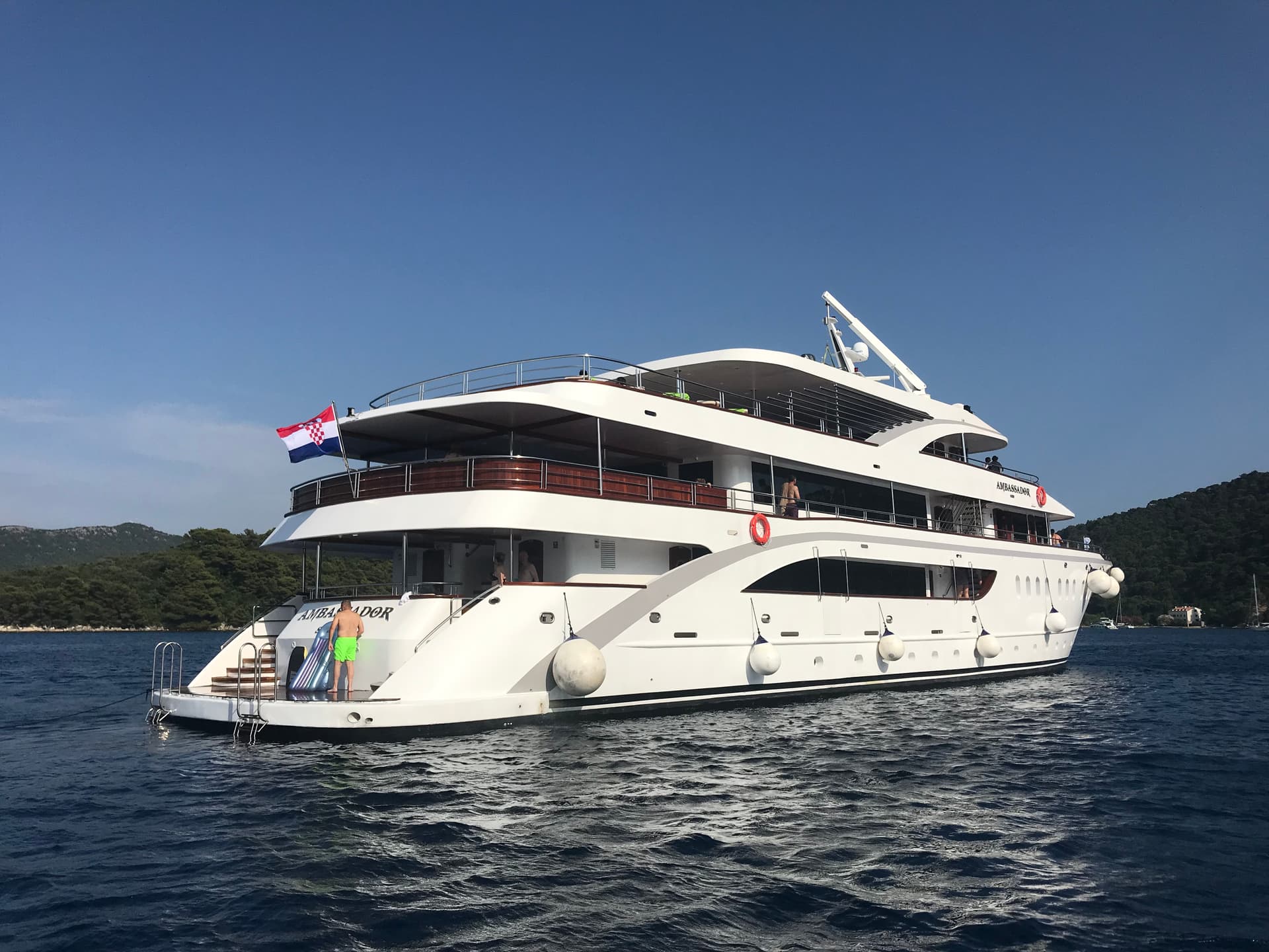 Large white yacht anchored on blue coastal water with wooded shores