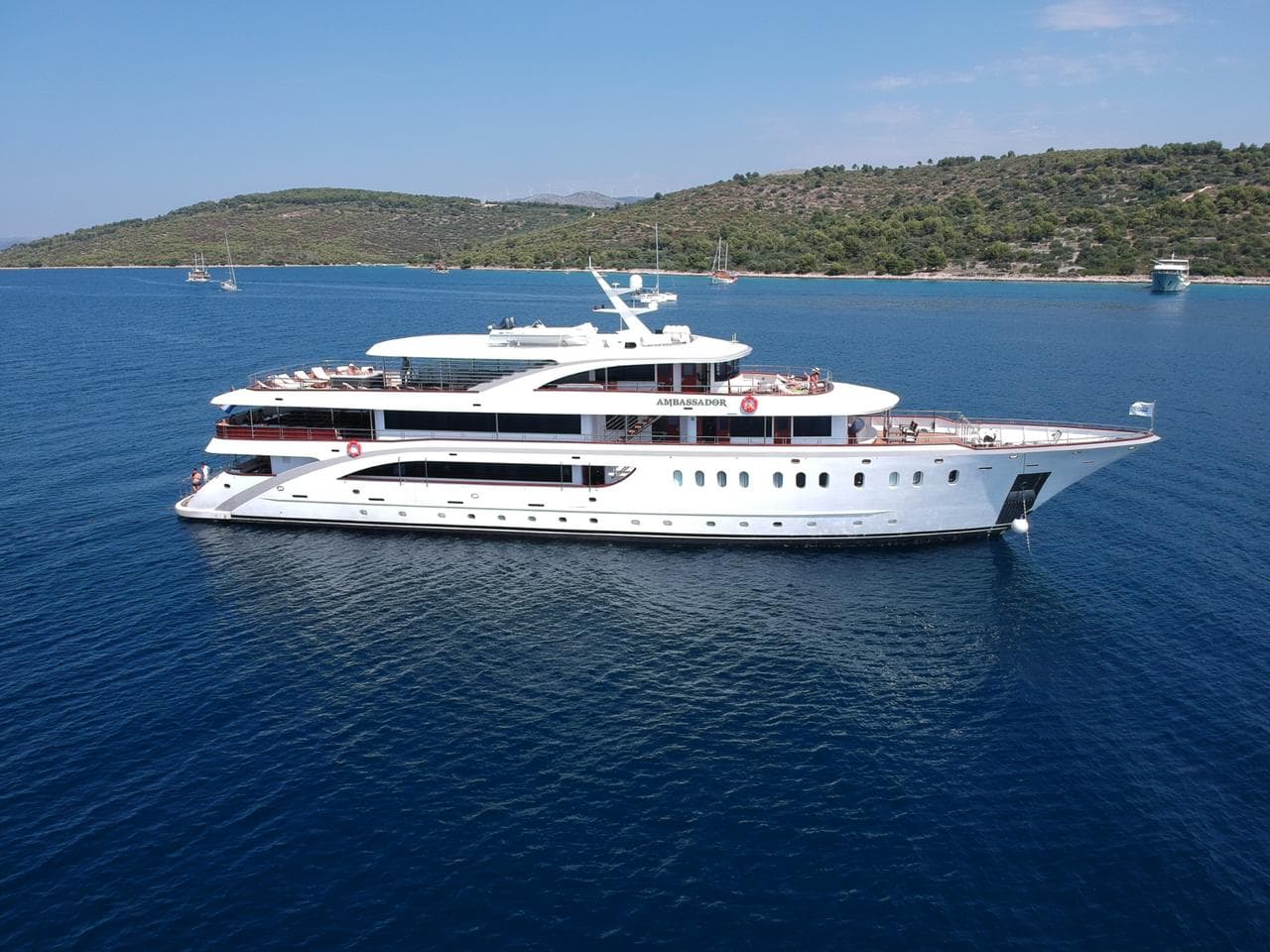 Large white Ambassador yacht anchored on calm blue coastal waters with green hills in background