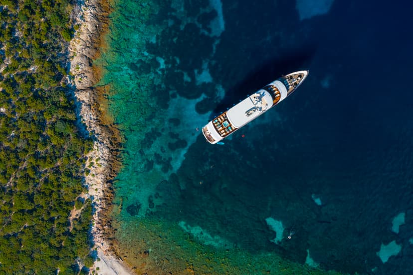 Yacht anchored in turquoise coastal waters near a rocky, forested shore