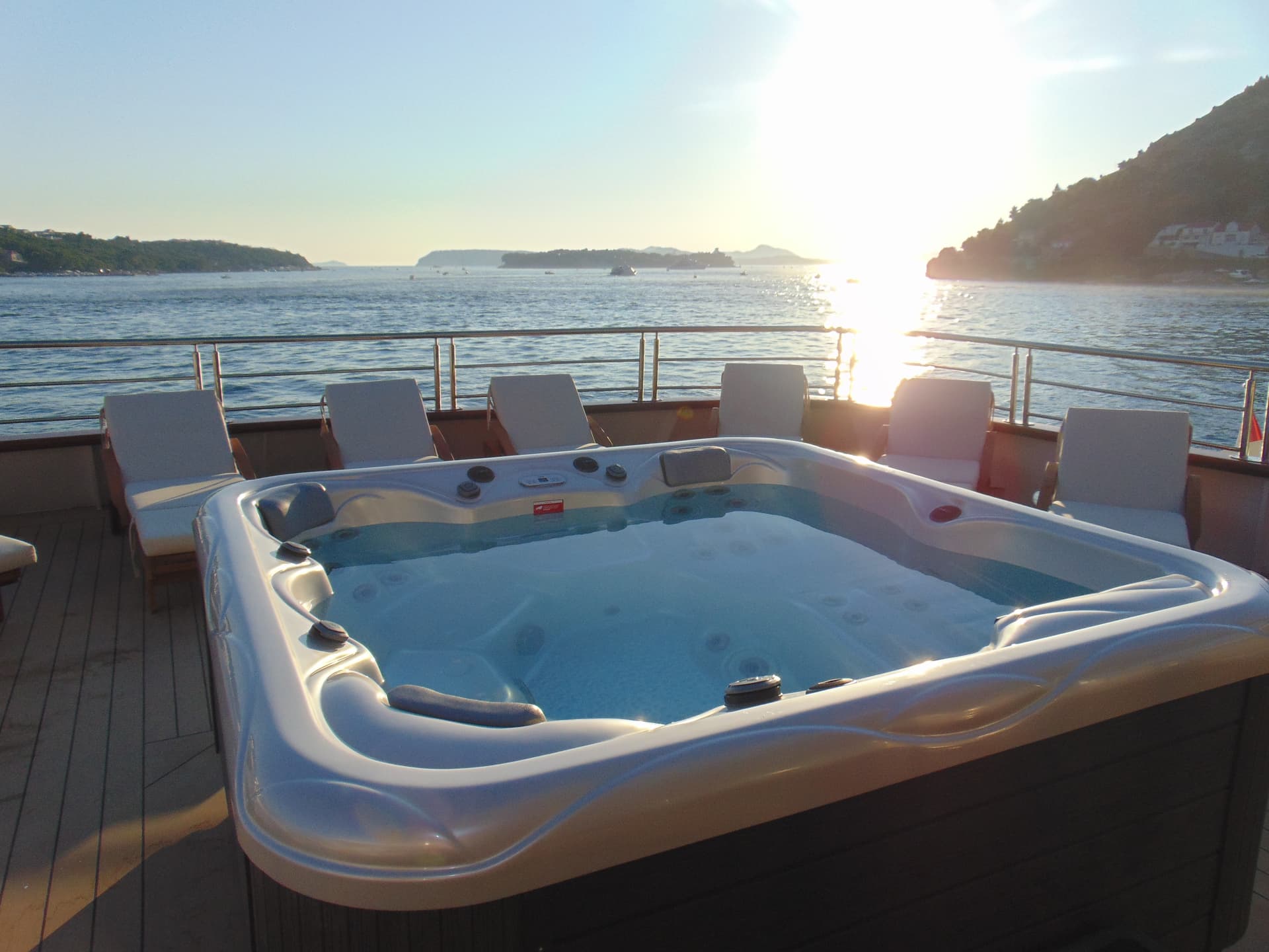 Hot tub on yacht deck with sun loungers overlooking coastal waters and islands at sunset