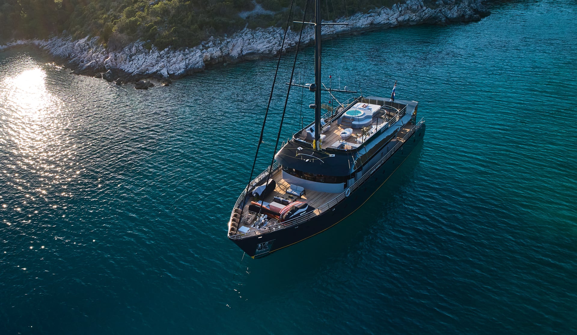 Luxury sailing yacht anchored near a rocky, vegetated coastline with sun reflecting on dark blue water.