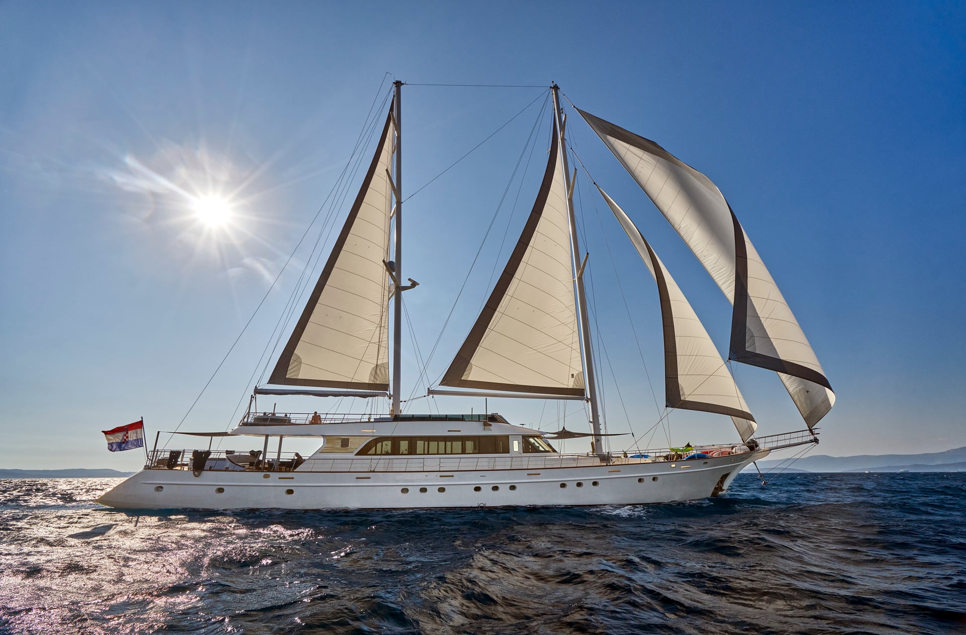 White yacht with sails up sailing on dark blue water under bright sun near distant mountains.