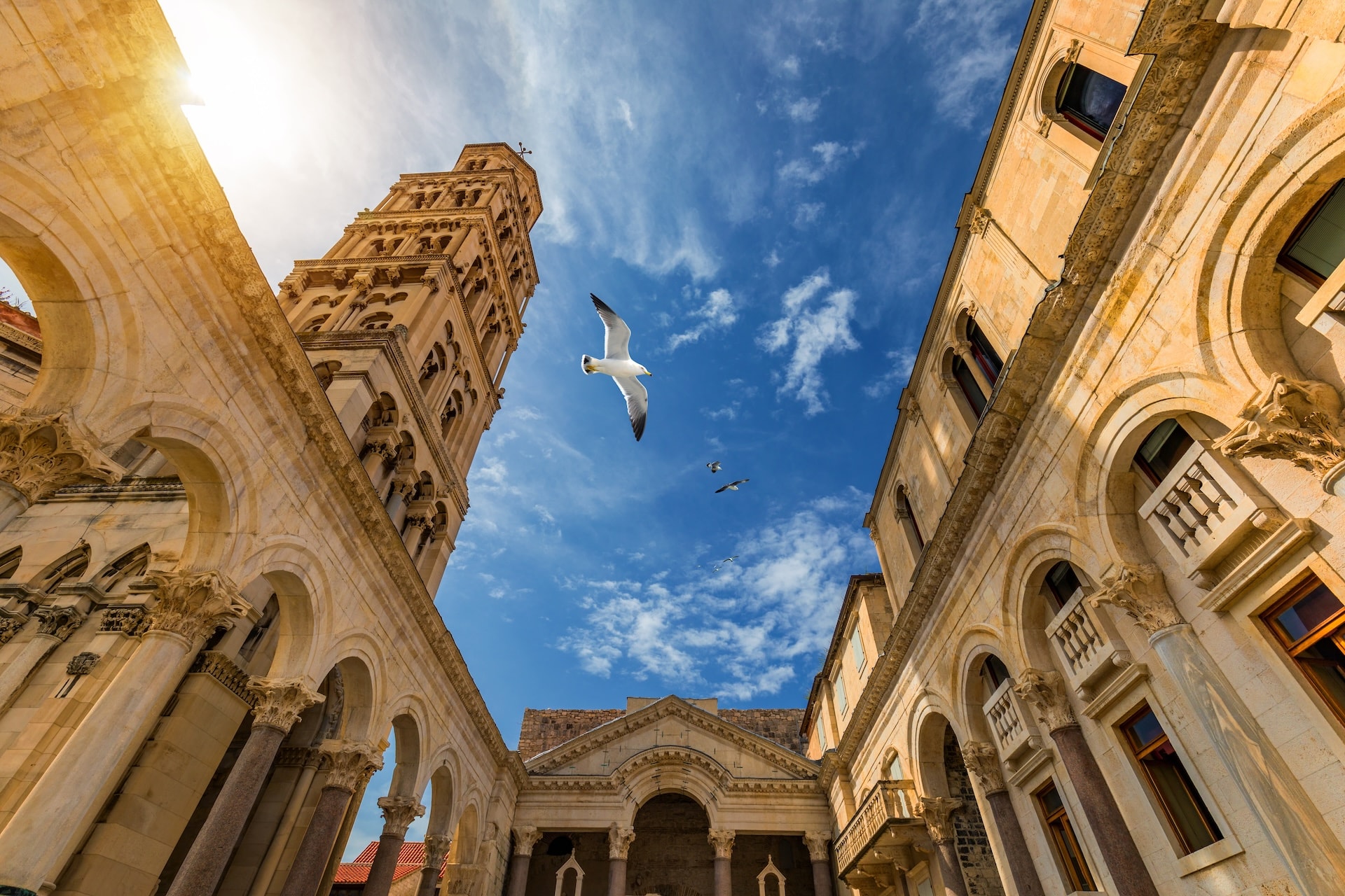 Bell tower and stone arches of Diocletian's Palace with seagulls flying against a bright blue sky.