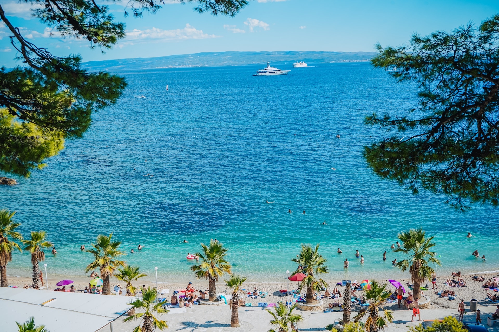 Yacht and ferry on blue sea near palm-lined beach with swimmers, viewed through tree branches.