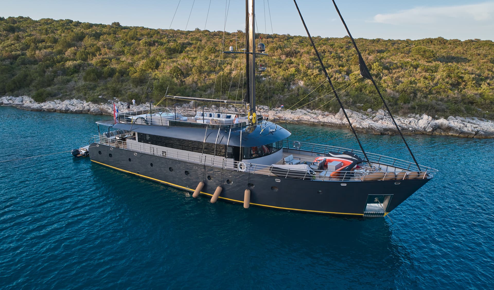 Large dark yacht anchored in deep blue water near a rocky, green coastline.