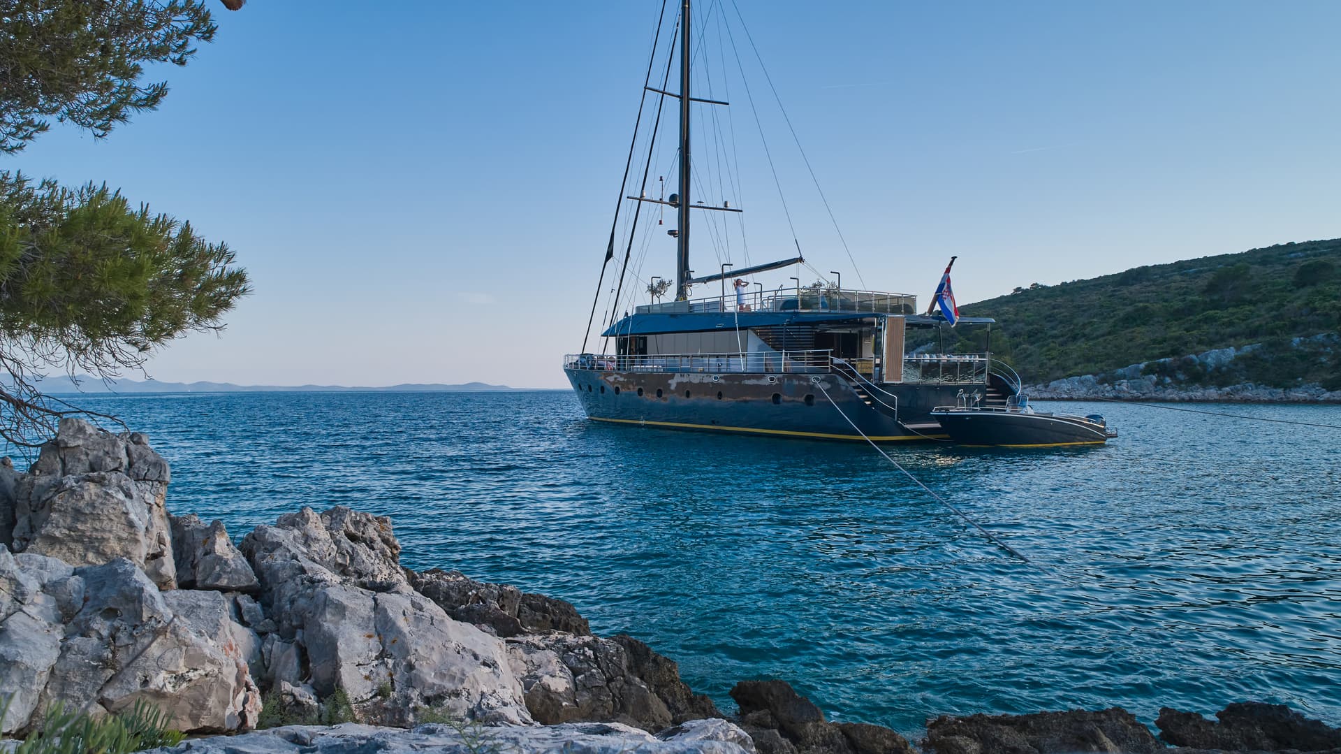 Large yacht anchored near rocky coastline with green hills under clear blue sky