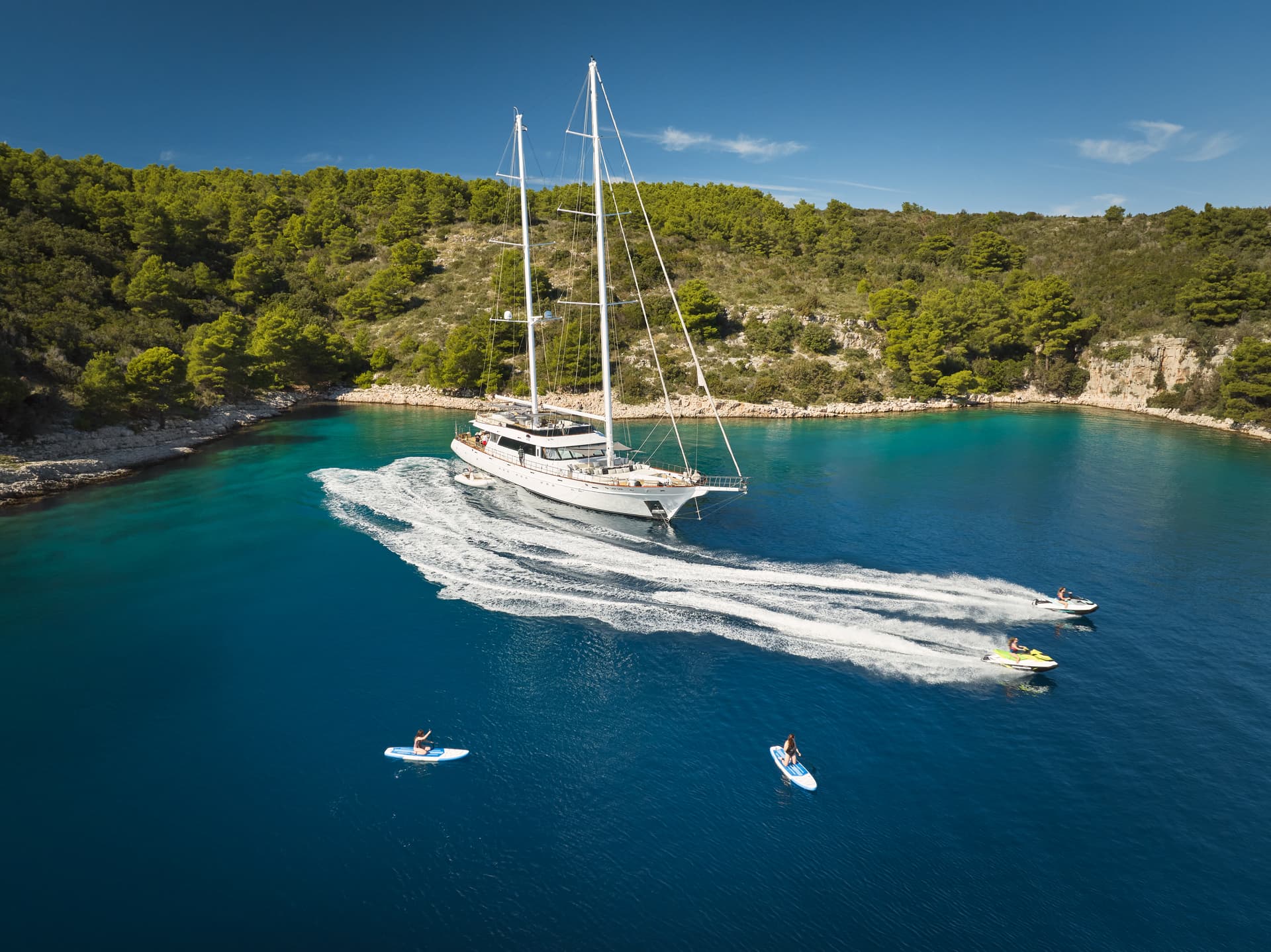 Large white sailboat in turquoise cove with jet skiing and paddle boarding activities.