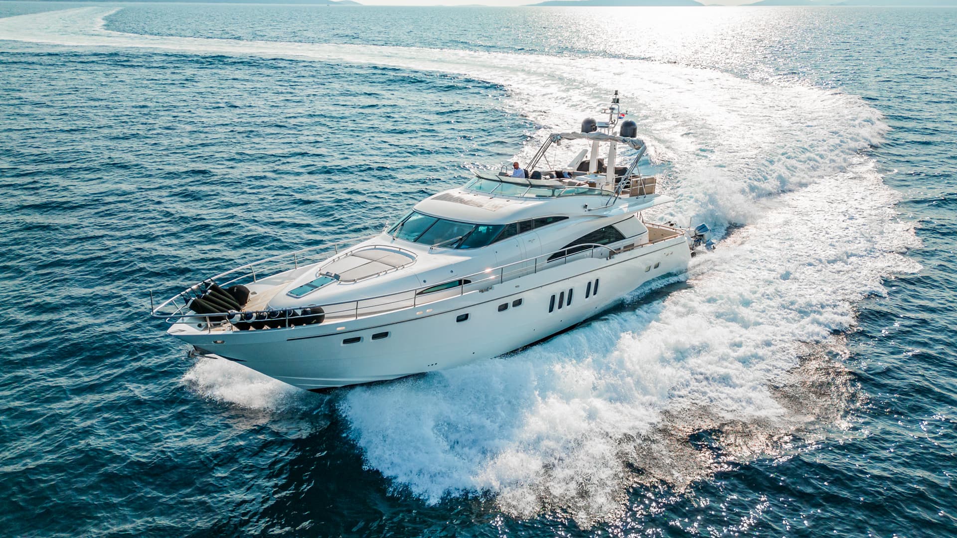 White motor yacht speeding across blue water, leaving a large wake toward distant land.