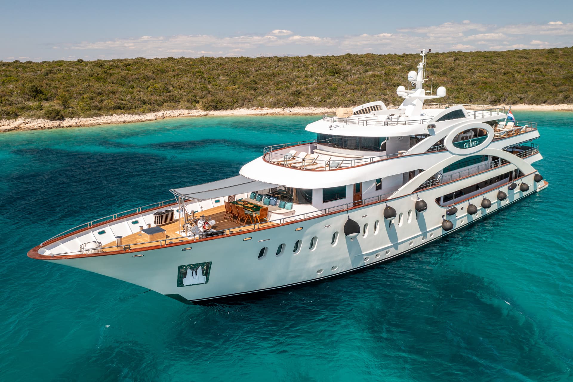 Large white yacht anchored in turquoise coastal waters near a densely vegetated shoreline.