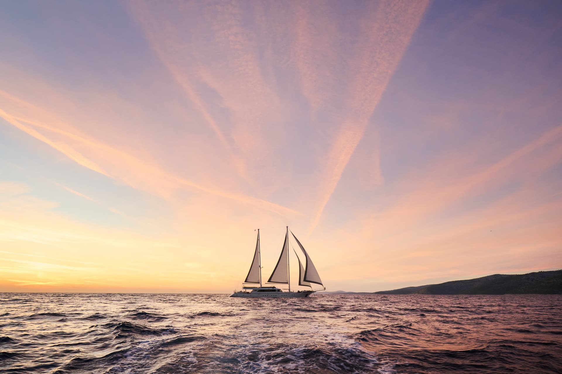 Large sailboat sailing on choppy sea water near a hilly coastline at sunset.