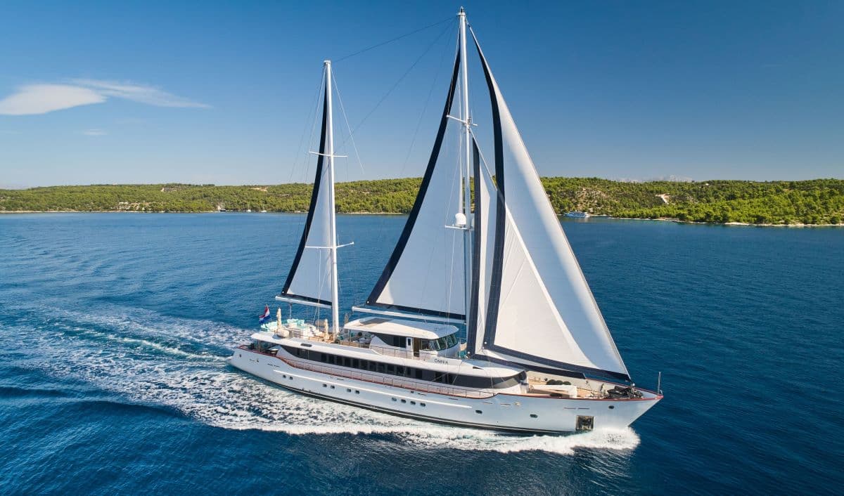 Large white sailing yacht named OMNIA cruising on deep blue sea near forested coastline.