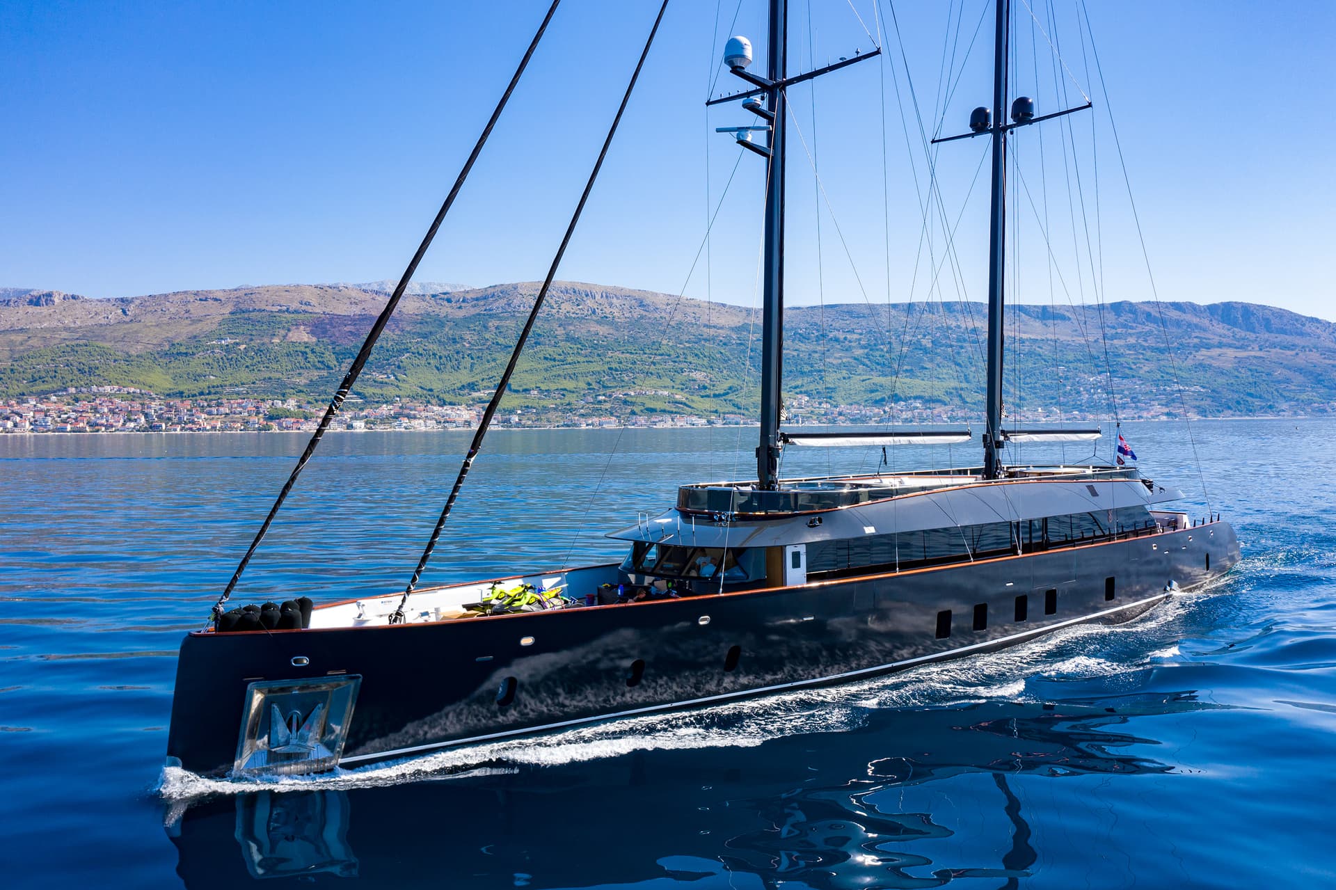 Large black sailing yacht cruising on blue water near a coastal town and hills.