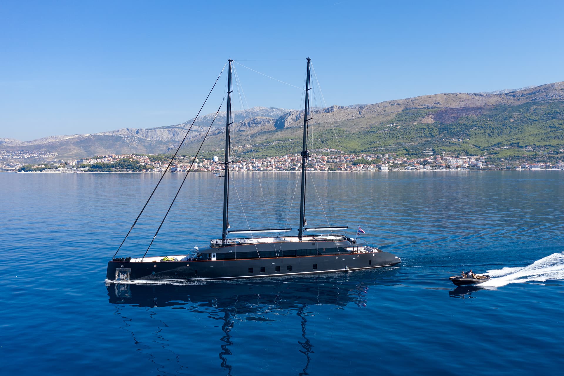 Large black sailboat and tender boat on deep blue sea near coastal town and mountains.
