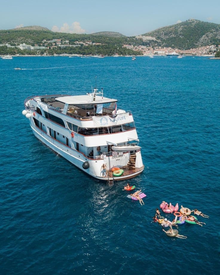 Large white boat anchored in blue sea with people swimming on floats near hilly coastline.