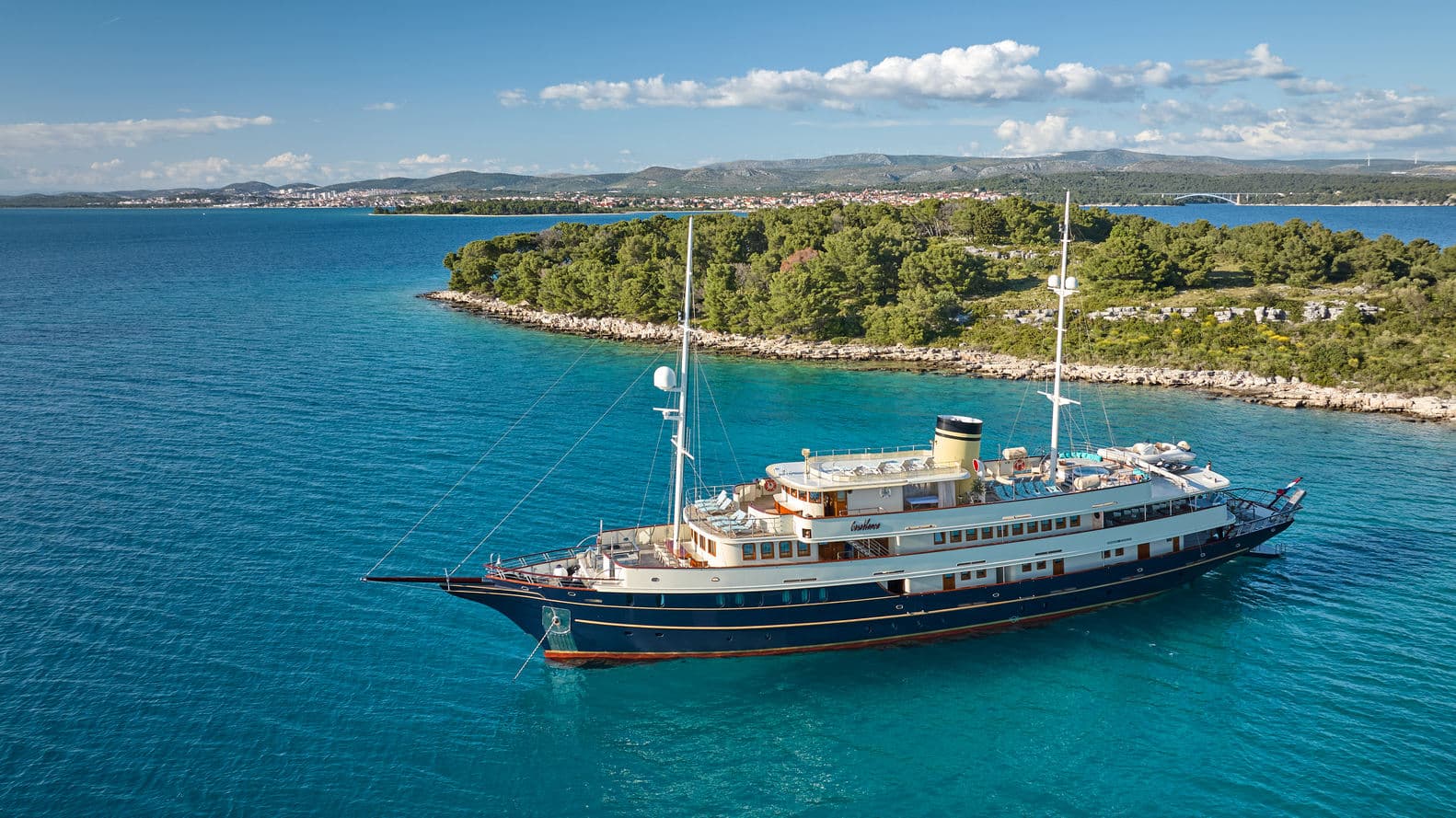 Luxury yacht anchored in turquoise coastal waters near a wooded island with a distant city.