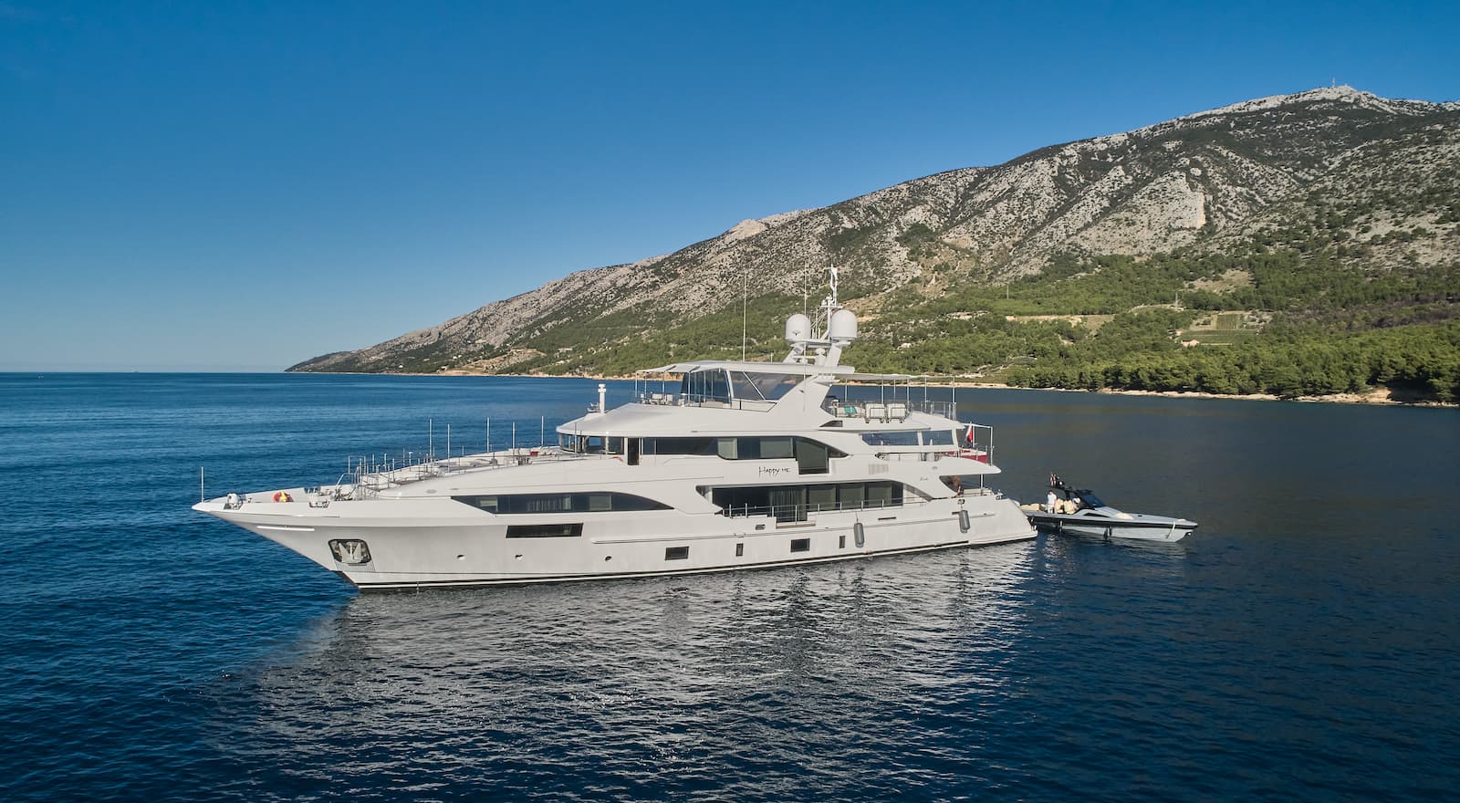 Large white motor yacht anchored near a steep, forested coastline under a clear blue sky.