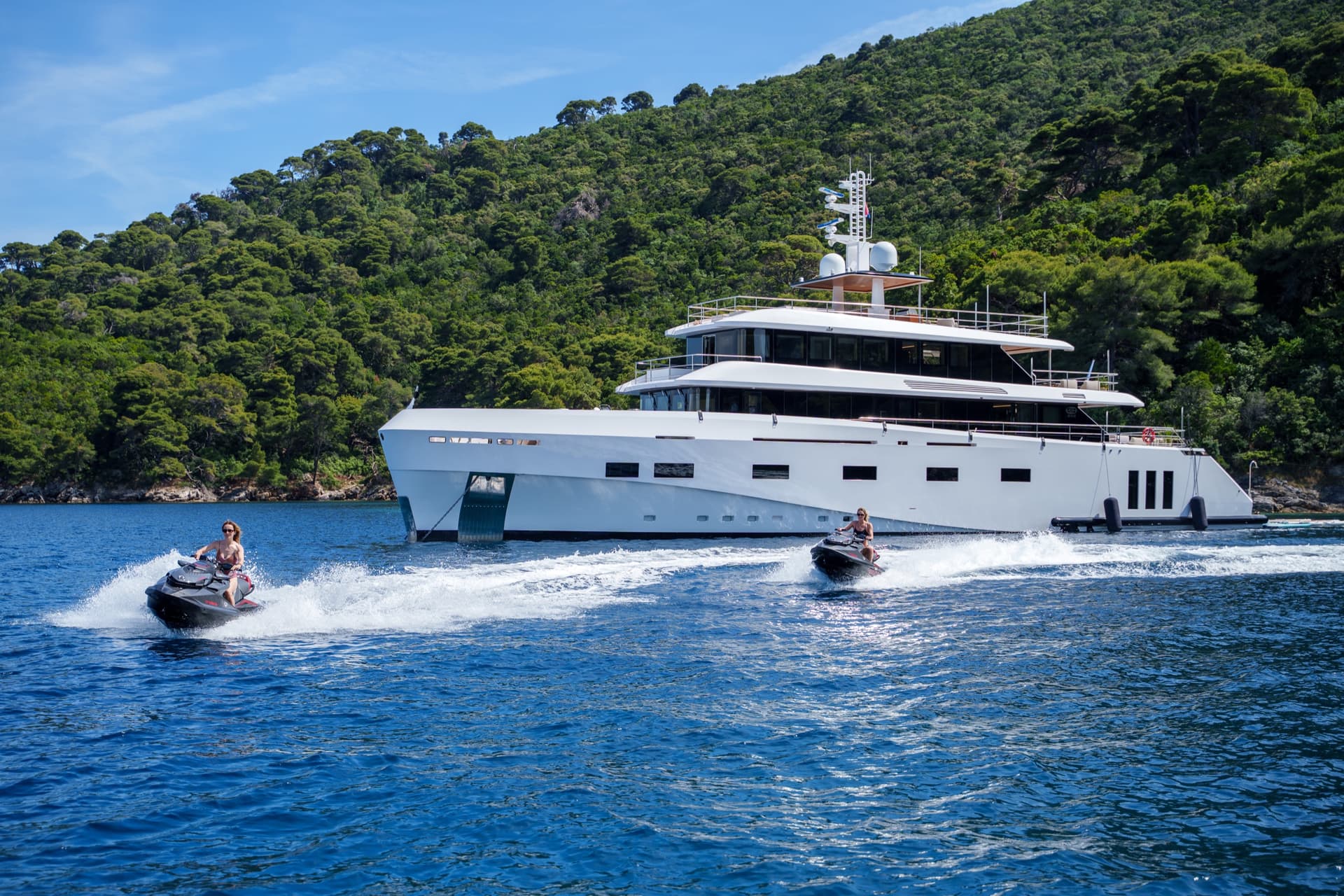 Large white luxury yacht anchored in coastal waters with people riding jet skis