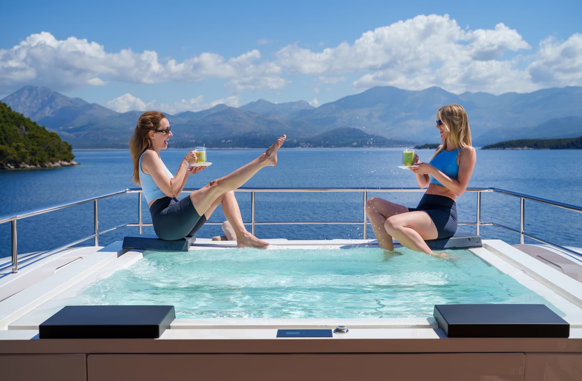 Two women drinking beverages by a yacht jacuzzi with mountain and sea views