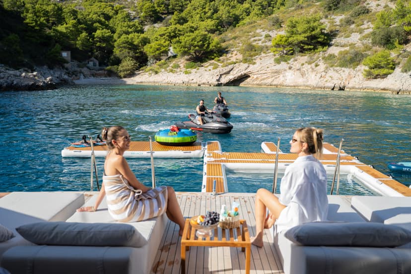 People relaxing on a yacht platform with floating beach toys and jet skis in a coastal bay