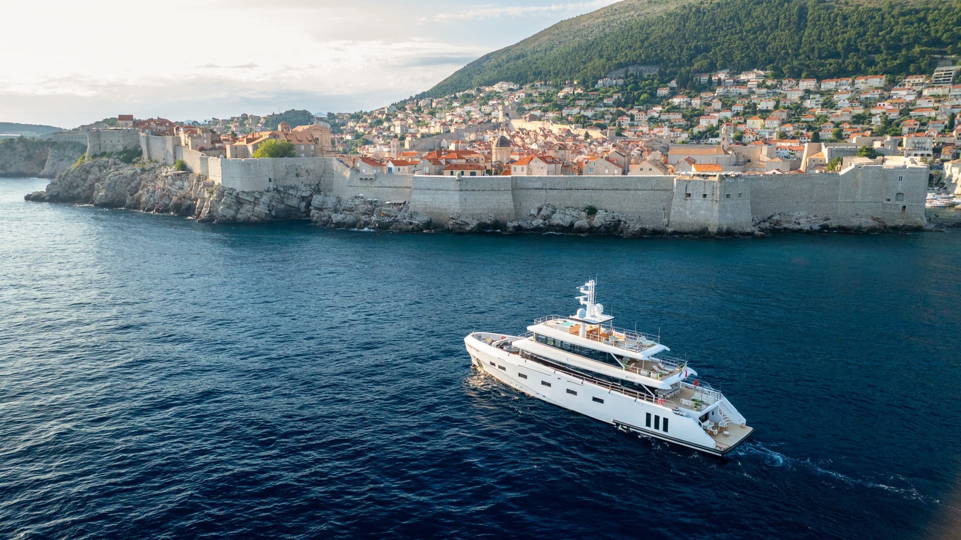 Yacht cruising on blue coastal waters near a historic walled city with terracotta roofs