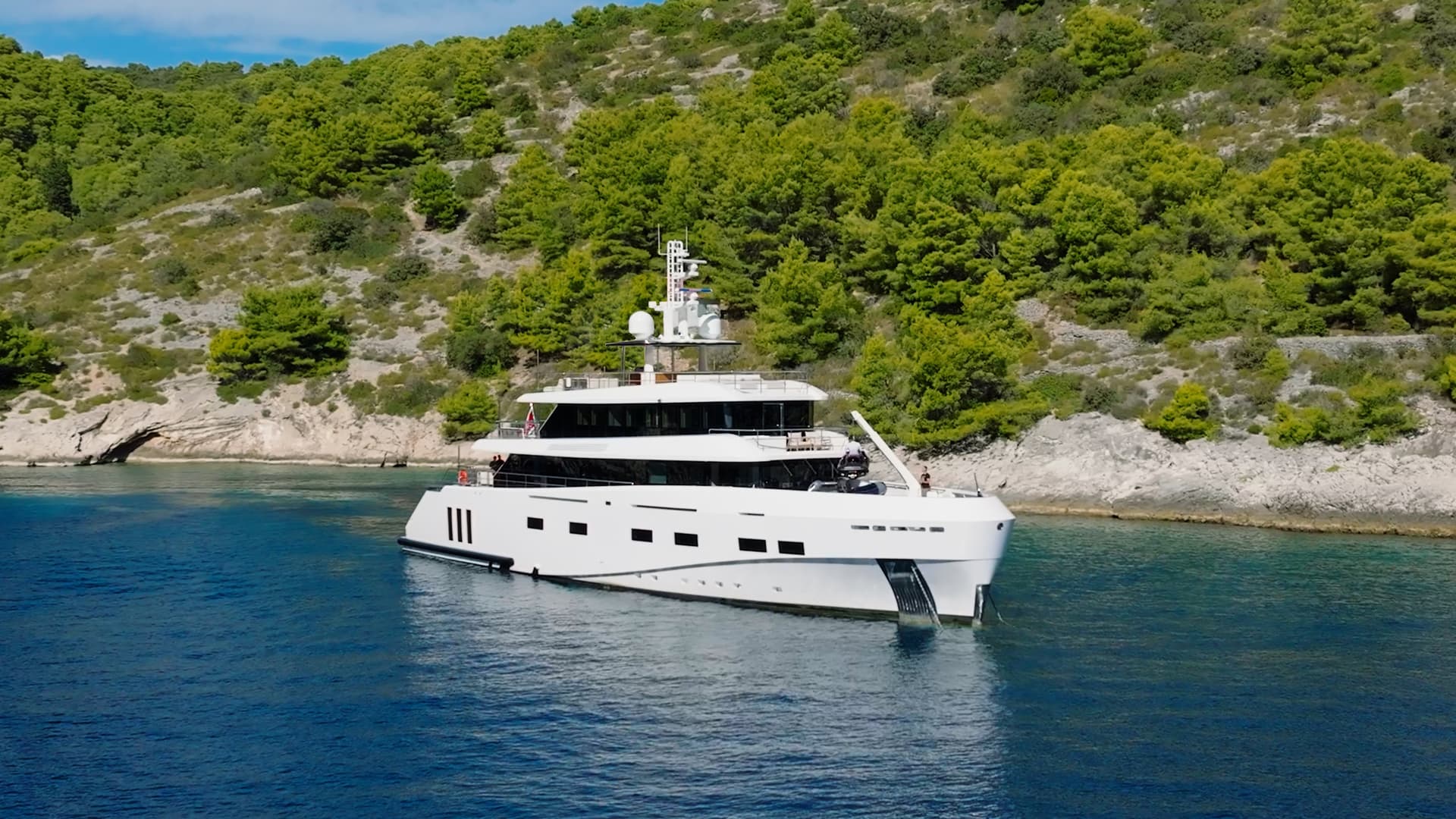White luxury yacht anchored in coastal waters near green wooded hills