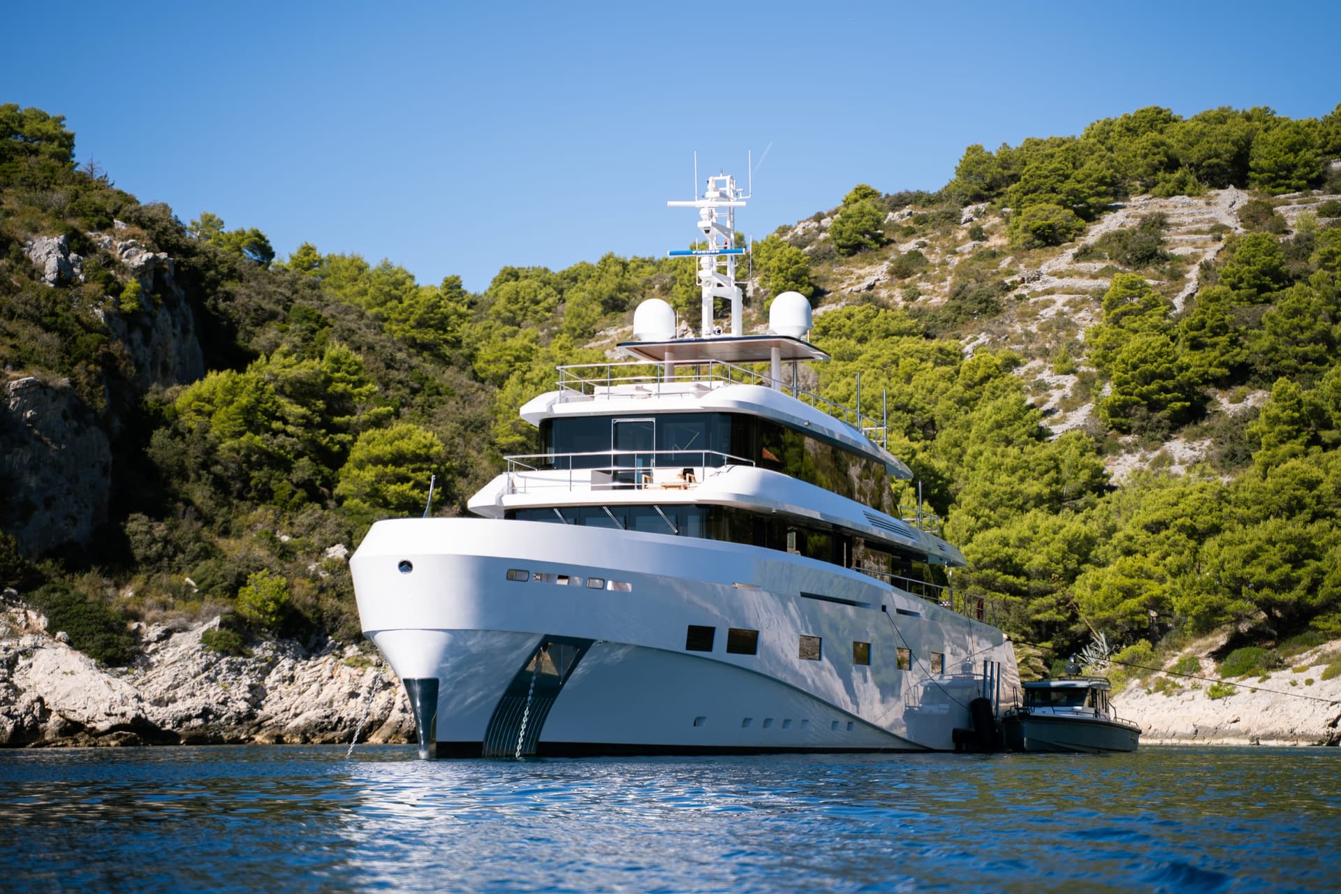 Large white luxury yacht anchored in a calm coastal bay with green forested hills